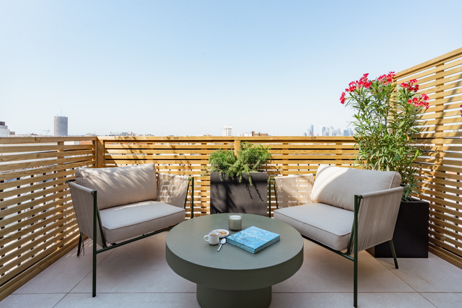 Deux fauteuils, une table basse et des lauriers fleuris sur la terrasse privée de l’Échappée Terrasse, avec les toits en arrière-plan